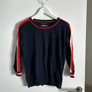 Mörkblå tröja från Tommy Hilfiger - Säljer en snygg mörkblå tröja från Tommy Hilfiger. Den har röda och vita ränder längs ärmarna och runt halsringningen. Perfekt för en casual look och passar bra till både jeans och chinos. Tröjan är i bra skick och redo för en ny ägare!