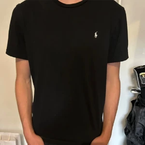 Svart t-shirt från Ralph Lauren - Säljer en klassisk svart t-shirt från Ralph Lauren med den ikoniska loggan broderad på bröstet. T-shirten är kortärmad och perfekt för en stilren look. Passar både till vardags och fest!
