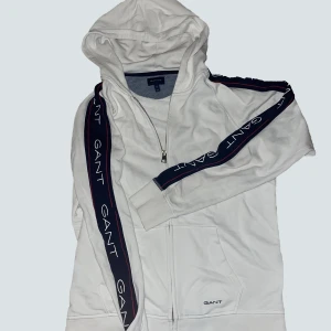 Vit hoodie från GANT - Säljer en snygg vit hoodie från GANT i storlek M. Den har en dragkedja framtill och GANT-loggan tryckt längs ärmarna. Perfekt för en casual look eller när du vill hålla det stilrent. Superbekväm och passar till det mesta! 💫 