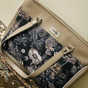 Beige och blommig väska från Sweet Bags - Säljer en superfin väska från Sweet Bags! Den är beige med ett snyggt blommigt mönster i svart och grått. Väskan har en dragkedja upptill och är perfekt för både vardag och lite finare tillfällen. Insidan är rymlig med flera fack för att organisera dina grejer. Perfekt för höst och vår!