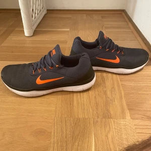 Nike träningsskor med orange detaljer - Säljer ett par snygga Nike träningsskor i mörkgrått med coola orange detaljer. De har en vit sula som ger bra dämpning och komfort. Perfekta för träning eller en aktiv vardag. Skorna har snörning för en bra passform och är i nyskick. 