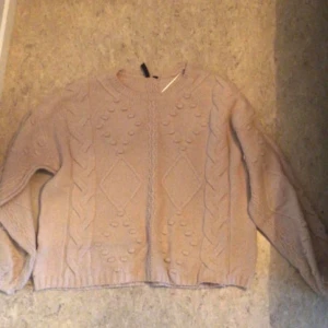 Beige stickad tröja - Säljer en supermysig beige stickad tröja med ett snyggt flätmönster och små bollar som detaljer. Perfekt för kyliga dagar och passar till allt! Tröjan har långa ärmar och en bekväm passform.