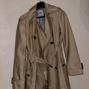 Trenchcoat/Kappa Zara Women - En superfin och kostym vibbar kappa från Zara Women i storlek S. Fickor, skärp och en stor luva. Fina knappar och väldigt detaljerad jacka. Vattentätt material och väldigt varm. I väldigt bra skick, inga skador/fläckar eller hål.