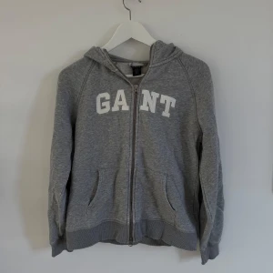 Grå hoodie från GANT - Säljer en snygg och bekväm grå hoodie från GANT. Den har en dragkedja framtill och en stor logga på bröstet. Perfekt för en avslappnad stil och passar till alla årstider. Den är i bra skick !