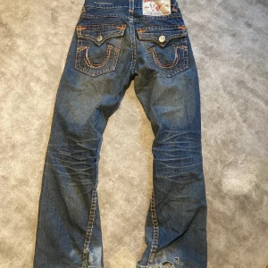 Blå jeans med detaljerad bakficka - Säljer ett par snygga blå jeans från True Religion. De har en cool design med detaljerade bakfickor och orange sömmar som verkligen poppar. Perfekta för en casual look eller en utekväll. De är i bra skick och redo för en ny ägare!