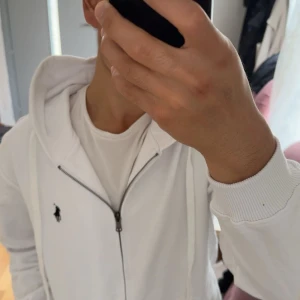 Vit hoodie från Polo Ralph Lauren - Säljer en snygg vit hoodie från Polo Ralph Lauren i storlek M. Den har en klassisk design med dragkedja och en broderad logga på bröstet. Hoodien är i nyskick och perfekt för både vår och höst. Den är gjord av mjukt bomullsmaterial och har en bekväm passform.