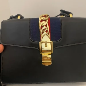 Svart axelväska  - Elegant svart axelväska  med en iögonfallande guldfärgad låsdetalj och kedja. Väskan har en röd och blå rand som ger en klassisk touch. Perfekt för att lyfta vilken outfit som helst. Har lite repor på guld detaljer men den syns inte så mycket