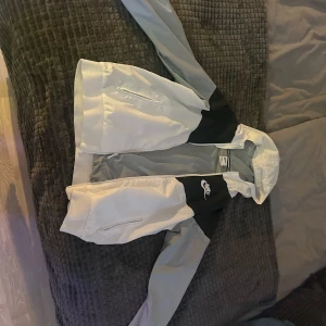 Svart och vit vindjacka från Nike - Säljer en stilren svart och vit vindjacka från Nike. Jackan har dragkedja och är perfekt för blåsiga dagar. Den har långa ärmar och en klassisk design med Nike-logga på bröstet.