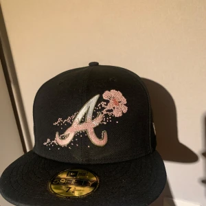 Svart keps med broderad logga och blomma - Snygg svart keps från New Era med en broderad logga och rosa blommor på framsidan. Kepsen har en platt skärm och är en del av 59FIFTY-serien. Perfekt för att ge din outfit en cool touch. Knappt använd, jag skulle gissa på att jag har haft utanför mitt hem cirka 3-4 gånger då den är för stor