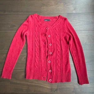 Röd kofta från Vero Moda - Säljer en snygg röd kofta från Vero Moda med knappar framtill. Den har ett klassiskt flätmönster och ribbade kanter. Perfekt för kyliga dagar!