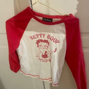 Vit och röd långärmad tröja med Betty Boop-tryck - Säljer en cool vit och röd långärmad tröja med Betty Boop-tryck. Tröjan har en klassisk raglanstil med kontrasterande röda ärmar och en tryckt design på framsidan. Perfekt för dig som gillar retrostil!