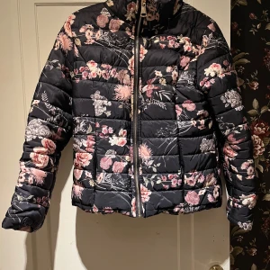 Blommig pufferjacka - Snygg svart pufferjacka med blommigt mönster i rosa och vita nyanser. Jackan har en dragkedja framtill och långa ärmar. Perfekt för att ge en färgglad touch till din outfit. Saknas dock en luva som ska vara till, se dragkedja på baksidan. 