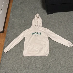 Vit hoodie från Björn Borg - Säljer en stilren vit hoodie från Björn Borg med texten 'BORG SINCE 1956' i grönt på bröstet. Tröjan har en klassisk känguruficka och justerbar huva med snören. Perfekt för en avslappnad stil.