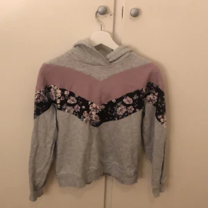 Grå hoodie med blommigt mönster från Kappal - Säljer en snygg grå hoodie från Kappal med en unik design. Tröjan har ett blommigt mönster i svart och rosa på ärmarna och en rosa detalj över bröstet. Perfekt för en avslappnad stil.