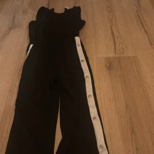 Svarta byxor med knappar från Lindex - Snygga svarta byxor från Lindex med vita knappar längs sidorna. Perfekta för en cool och avslappnad stil. Byxorna har en elastisk midja för extra komfort.