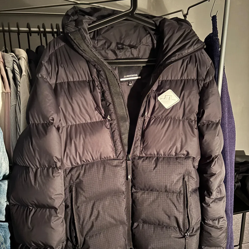 Snygg svart dunjacka från J.Lindeberg med huva och dragkedja. Jackan har ett stilrent märke på bröstet och är perfekt för kyligare väder. Den quiltade designen ger extra värme och komfort.. Takit.