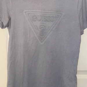 Grå t-shirt från Guess - Snygg grå t-shirt från Guess med det ikoniska triangellogotypen i samma färg på bröstet. Perfekt för en stilren och avslappnad look.
