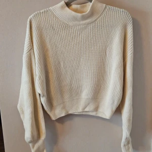 Beige stickad tröja från NA-KD - Säljer en stilren beige stickad tröja från NA-KD. Tröjan har en hög krage och långa ärmar, perfekt för kyligare dagar. Den är croppad och har en ribbad struktur som ger en snygg look. Tröjan är aldrig använd.