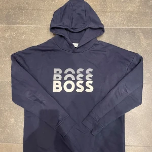 Mörkblå hoodie från Boss - Säljer en stilren mörkblå hoodie från Boss med stor logga på framsidan. Tröjan har långa ärmar och en klassisk huva. Perfekt för en avslappnad stil.