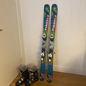 Twintipskidor och pjäxor - Säljer ett par svarta skidpjäxor från Dalbello i pjäxstorlek 25,5 med justerbara spännen och snygga vita detaljer. Perfekta för vinterns skidäventyr. Passar bra med Firefly skidor. Säljer ett par Firefly twintipskidor i längden 145 cm. Skidorna har en stött kant på tippen men påverkar inte funktionen. 