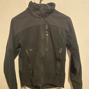 Svart fleecejacka från Haglöfs - Säljer en svart fleecejacka från Haglöfs med dragkedja och hög krage. Jackan har praktiska fickor med dragkedjor och är perfekt för kyliga dagar. Den är designad för att hålla dig varm och bekväm. Säljer denna Haglöfs jacka då den har blivit för liten för mig. 