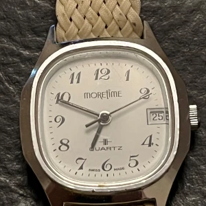 Klocka från Moretime - Stilren klocka från Moretime med vit urtavla och datumvisning. Boetten är i silverfärgad metall och har en kvadratisk form. Klockan har ett flätat beige armband som ger en unik touch. Swiss made och vattentålig med quartz-urverk.