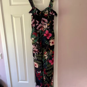 Blommig byxdress med volangdetalj - Snygg byxdress med blommigt mönster i rosa, grönt och rött på svart bakgrund. Den har volangdetaljer vid axelbanden och en smickrande passform. Perfekt för fest!