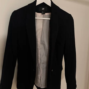 Svart kavaj från H&M - Snygg svart kavaj från H&M i storlek S. Perfekt för en stilren look med klassisk design och långa ärmar. Kavajen har en enkel knäppning framtill och är fodrad med ett ljust tyg.