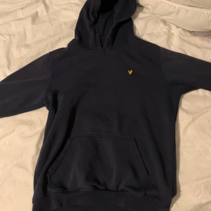 Mörkblå hoodie från Lyle & Scott - Säljer en stilren mörkblå hoodie från Lyle & Scott med en liten gul logga på bröstet. Tröjan har en klassisk känguruficka och en bekväm huva. Perfekt för en avslappnad stil.