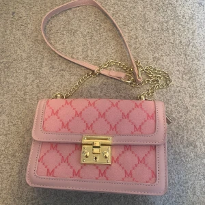 Rosa axelväska med guldkedja - Snygg rosa axelväska med guldkedja och mönstrad framsida. Väskan har ett elegant lås i guld och en praktisk innerficka med dragkedja. Perfekt för att ge en färgklick till din outfit.