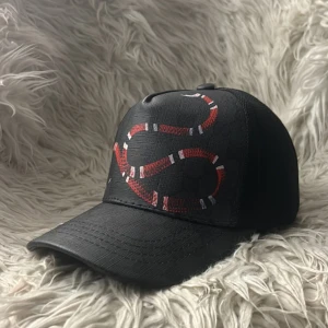Svart keps med röd orm - Snygg svart keps med en cool röd orm broderad på framsidan. Kepsen har en justerbar rem baktill med röd och blå detalj. Perfekt för att ge din outfit en edgy touch.