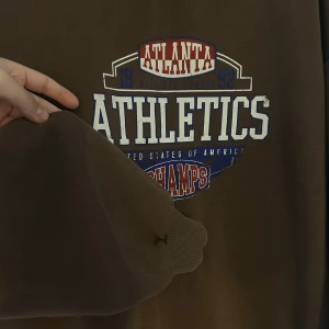 Brun sweatshirt med tryck 'Atlanta Athletics' - Säljer en brun sweatshirt med trycket 'Atlanta Athletics' i vitt, rött och blått. Tröjan har långa ärmar och en rund halsringning. Perfekt för en avslappnad stil. Den har storlek XL , men passar som M/S 