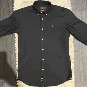Svart skjorta från Morris - Snygg svart skjorta från Morris med klassisk krage och vita knappar. Skjortan har en broderad detalj på bröstet och är perfekt för en stilren look. Passar bra till både vardag och fest. Storlek S men mer som en M