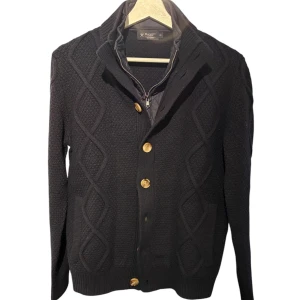 Navy stickad kofta från Hackett - Navy svart stickad kofta från Hackett med knappar och dragkedja. Den har ett elegant mönster och är perfekt för kyligare dagar. Koftan är tillverkad i ull och har en stilren design med ribbade detaljer.