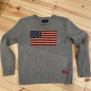 Grå stickad tröja från Polo Ralph Lauren - Säljer en stilren grå stickad tröja från Polo Ralph Lauren med ett broderat amerikanskt flaggmotiv på framsidan. Tröjan har långa ärmar och ribbstickade muddar vid ärmslut och nederkant. Perfekt för en avslappnad och klassisk look.