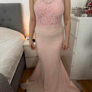 Ljusrosa långklänning med spetsdetaljer - Elegant ljusrosa långklänning med spetsöverdel och hög hals. Klänningen har en figurnära passform och en vacker spetsdetalj på ryggen. Perfekt för formella tillfällen.