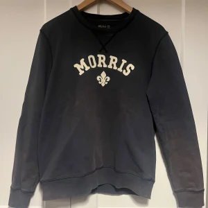Svart sweatshirt från Morris - Snygg svart sweatshirt från Morris med vit text och emblem på bröstet. Tröjan har långa ärmar och en klassisk rund halsringning. Perfekt för en avslappnad stil.