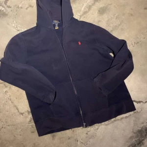 Mörkblå hoodie från Ralph Lauren - Säljer en snygg mörkblå hoodie från Ralph Lauren med dragkedja och det klassiska röda logotypbroderiet på bröstet. Perfekt för en avslappnad stil. Priset är inte hugget i sten!