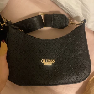 Svart axelväska från Guess - Snygg svart axelväska från Guess med präglat mönster och justerbar axelrem. Väskan har en dragkedja upptill och guldiga detaljer som ger en lyxig känsla. Perfekt för att bära dina nödvändigheter med stil. Aldrig använd💓💓