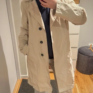 Beige trenchcoat - Snygg beige trenchcoat med knappar framtill. Perfekt för vår och höst. Klassisk design och normal passform.