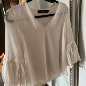 Vit blus  - Elegant vit blus från Zara med v-ringning och långa ärmar. Ärmarna har vackra volangdetaljer som ger en romantisk touch. Perfekt för både vardag och speciella tillfällen.