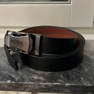 Svart läderbälte från Hugo Boss - Snyggt svart läderbälte från Hugo Boss med en stilren silverfärgad spänne. Perfekt för att ge en elegant touch till din outfit. Bältet har en klassisk design som passar till många olika stila.