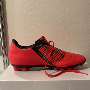 Röda Nike fotbollsskor - Snygga röda Nike fotbollsskor med svarta detaljer och snörning. Skorna har en slitstark sula med dobbar för optimalt grepp på planen. Perfekta för fotbollsträning och matcher.