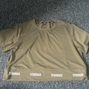 Olivgrön t-shirt med text - Snygg olivgrön t-shirt med texten 'STRONGER' tryckt flera gånger längs nederkanten. Perfekt för en avslappnad stil eller träning. Kortärmad design med rund hals.