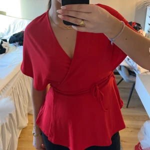 Röd omlottopp - Snygg röd omlottopp med korta ärmar och knytband i midjan. Perfekt för en stilren look. Köpt på Plick tidigare och har aldrig använt den då den inte är min stil