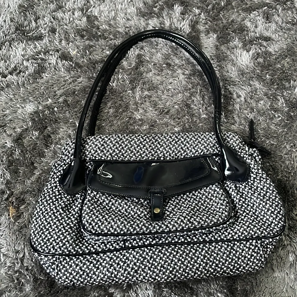 Snygg svartvit rutig handväska med svarta detaljer och blanka handtag. Väskan har en praktisk ficka framtill med knappstängning och dragkedja upptill. Perfekt för att ge din outfit en stilren touch.. Laukut & Käsilaukut.