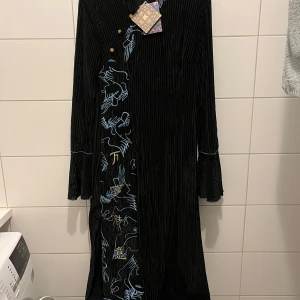 Svart långklänning Chi Pao nya  - Elegant svart långklänning med vackra broderade detaljer i blått och guld. Klänningen har en slits på sidan och långa ärmar. Perfekt för en stilfull look. Passar S-M