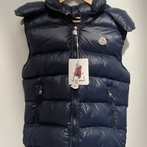 Moncler väst  - Säljer en snygg mörkblå dunväst från Moncler med dragkedja och huva. Västen har en quiltad design och Moncler-logga på bröstet. Perfekt för kyligare dagar. L passar även M eftersom att den är liten i storlek sitter fint på L och M. Den är helt ny och oanvänd , har en innreficka och är i bästa skick . Skriv för mer info! 