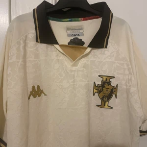 Riksfet vasco da gama fotbollströja - Snygg vit fotbollströja från Kappa med korta ärmar och ett stilrent mönster. Tröjan har en svart krage med gulddetaljer och ett emblem på bröstet. Perfekt för fotbollsfans som vill ha något unikt i garderoben.