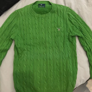 Grön stickad tröja från GANT - Säljer en snygg grön stickad tröja från GANT i premium bomull. Tröjan har ett klassiskt kabelstickat mönster och rund halsringning. Perfekt för en stilren look!
